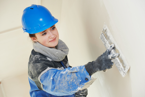 Person plastering a wall while wearing a blue construction hard hat, a gray scarf, a blue and black jacket, and black gloves.  The wall shows a partially plastered area. The background is plain and light-colored.